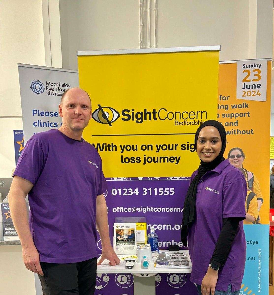 A man and a woman are standing either side of a table that displays Low Vision aids, a Sight Concern roller banner is behind them.. Both volunteers are wearing Sight Concern Bedfordshire t-shirts.